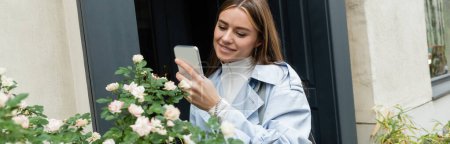 happy young woman in blue trench coat taking photo of green bush with blooming flowers on street in Vienna, banner 