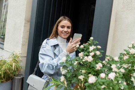 joyful young woman in blue trench coat taking photo of green bush with blooming flowers on street in Vienna 