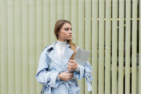 pretty woman in blue trench coat holding laptop while standing near window on street in Vienna 