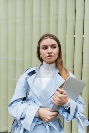 pretty freelancer in blue trench coat holding laptop while standing near window on street in Vienna 
