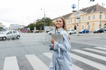Glückliche Frau im blauen Trenchcoat hält Laptop in der Hand und überquert Straße in Wien 