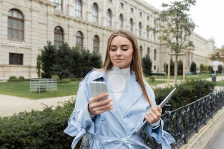 junge Frau in blauem Trenchcoat mit Laptop und Smartphone in der Nähe historischer Gebäude in Wien 