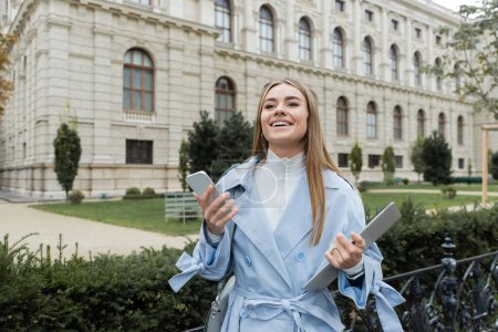 happy young woman in blue trench coat holding laptop and smartphone near historical building in Vienna 