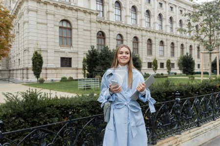 fröhliche Frau in blauem Trenchcoat mit Laptop und Smartphone in der Nähe historischer Gebäude in Wien 