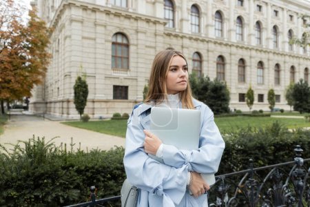 jolie jeune femme en trench coat bleu tenant ordinateur portable près du bâtiment historique à Vienne 