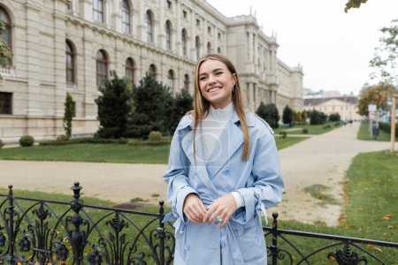 joyful young woman in blue trench coat standing near historical building in Vienna 