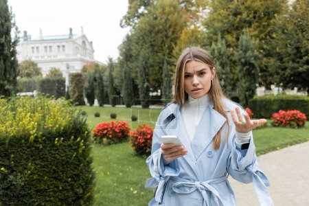 femme confus en trench coat bleu en utilisant smartphone tout en se tenant dans le parc vert à Vienne 
