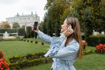 cheerful woman in blue trench coat taking selfie on mobile phone while standing in green park in Vienna 