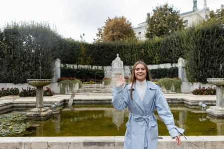 smiling young woman in blue trench coat waving hand near fountain in green park 