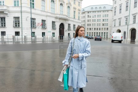 Téléchargez les photos : Jeune femme en trench coat bleu marchant avec des sacs à provisions tout en marchant près des bâtiments à Vienne - en image libre de droit