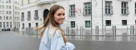 positive young woman in blue trench coat smiling on urban street in Vienna, banner 