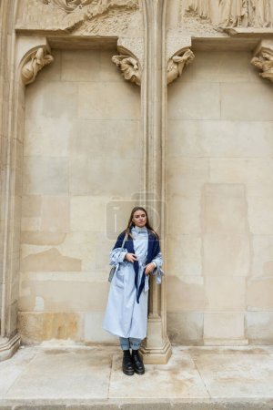 young woman with scarf on top of blue trench coat standing near historical building in Vienna 