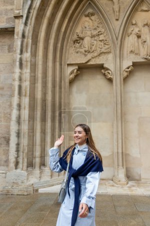 alegre joven con bufanda en la parte superior de la gabardina azul agitando la mano cerca del edificio histórico en Viena 