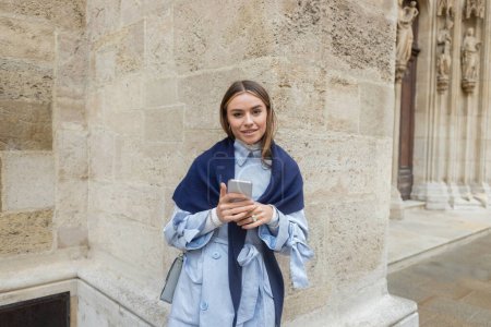 junge Frau mit marineblauem Schal über Trenchcoat und Handy in der Nähe historischer Gebäude in Wien 