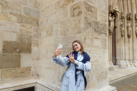 glückliche Frau mit Schal auf blauem Trenchcoat bei Videoanruf in der Nähe historischer Gebäude in Wien 