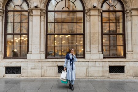 junge, stylische Frau mit Schal über blauem Trenchcoat mit Einkaufstaschen in der Nähe historischer Gebäude in Wien