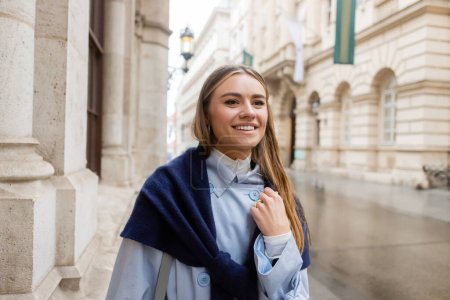 Téléchargez les photos : Femme heureuse et élégante avec écharpe sur le dessus de trench coat bleu souriant tout en regardant loin dans la rue à Vienne - en image libre de droit