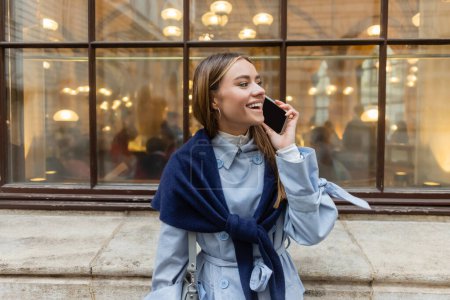 cheerful woman with scarf on blue trench coat talking on smartphone on street in Vienna