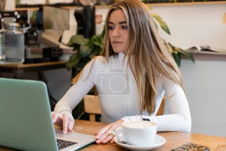 mujer encantadora en cuello alto usando el ordenador portátil cerca de la taza de capuchino en la cafetería 