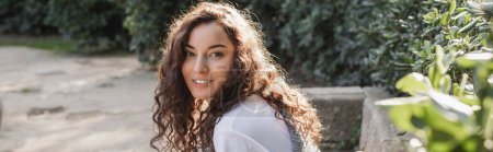 Téléchargez les photos : Femme frisée et jeune brune en t-shirt souriant à la caméra alors qu'elle était assise sur un banc de pierre et relaxante près de buissons verts dans un parc à Barcelone, Espagne, soleil, bannière - en image libre de droit