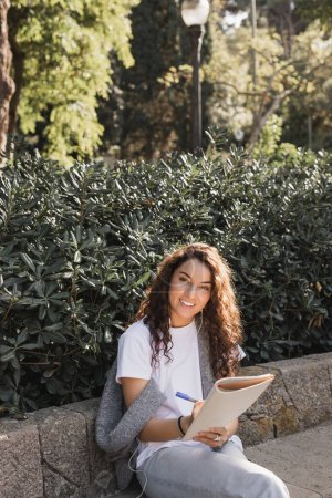 Mujer joven y rizada sonriente en ropa casual usando auriculares con cable, escribiendo en un cuaderno y mirando a la cámara mientras está sentada en un banco de piedra en el parque en Barcelona, España 