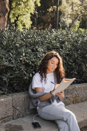 Young and curly brunette woman writing on notebook while listening music in wired earphones near smartphone on stone bench and green bushes in park at daytime in Barcelona, Spain 