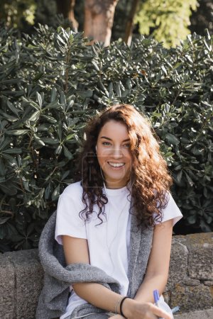 Portrait of cheerful and curly young woman in casual clothes using wired earphones and holding marker while sitting on stone bench in park at daytime in Barcelona, Spain 
