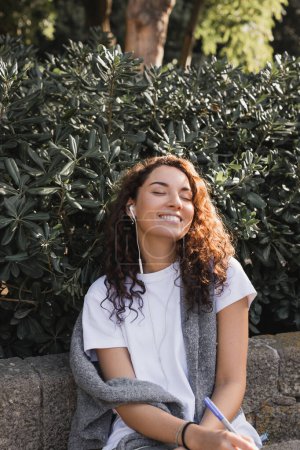 Carefree young and brunette woman with closed eyes in casual clothes listening music in wired earphones and smiling, holding marker while relaxing on stone bench in park in Barcelona, Spain 