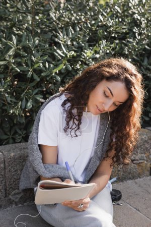 Jeune femme bouclée en vêtements décontractés écoutant de la musique dans des écouteurs filaires et écrivant sur un ordinateur portable tout en se relaxant sur un banc en pierre dans un parc à Barcelone, Espagne 