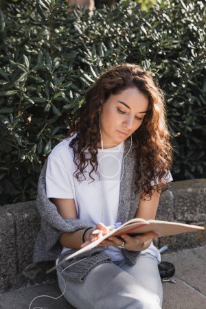 Joven mujer rizada y bonita con ropa casual y auriculares con cable mirando el cuaderno y sosteniendo el marcador mientras está sentada en un banco de piedra en el parque en Barcelona, España 