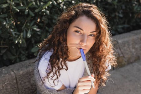 Portrait of young and thoughtful curly woman in t-shirt and sweater holding marker near lips and looking at camera while sitting on stone bench in park in Barcelona, Spain 