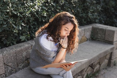 Mujer joven y rizada en ropa casual tocando los labios, sosteniendo el marcador y mirando el cuaderno mientras está sentada en un banco de piedra cerca de arbustos verdes en Barcelona, España 