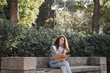 Mujer joven y rizada pensativa con ropa casual sosteniendo un cuaderno y un marcador cerca de los labios mientras está sentada cerca de dispositivos en un banco de piedra y plantas verdes en el parque durante el día en Barcelona, España 