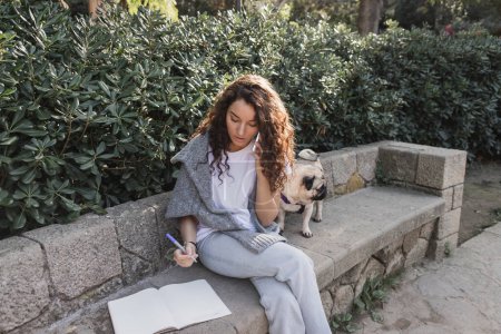 Mujer joven y rizada en suéter hablando en un smartphone y sosteniendo un marcador cerca de un cuaderno con espacio para copiar y un perrito en un banco de piedra cerca de arbustos verdes en el parque en Barcelona, España 