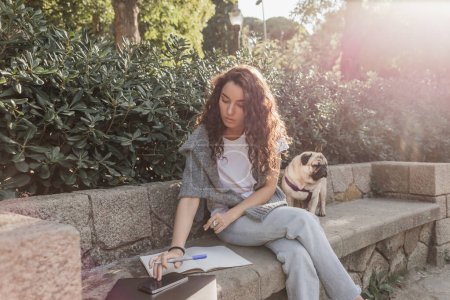 Hübsche und lockige junge Frau in lässiger Kleidung mit Smartphone in der Nähe von Laptop, Notizbuch und Mops-Hund, während sie auf einer steinernen Bank im grünen Park in Barcelona sitzt, Spanien, freiberuflich 