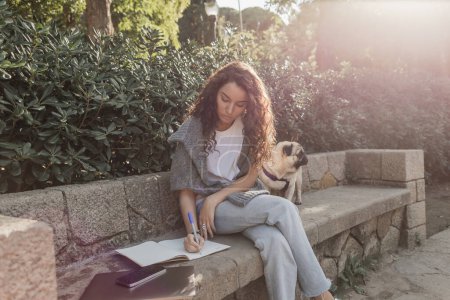 Lockenlockige junge Frau in lässiger Kleidung, die auf einem Notizbuch schreibt, während sie in der Nähe von Geräten und Mops-Hund auf einer Steinbank und grünen Büschen in einem Park in Barcelona sitzt, Spanien, freiberuflich 