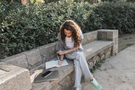 Junge und lockige Frau in warmem Pullover auf den Schultern, die auf einem Notizbuch neben Laptop und Smartphone mit leerem Bildschirm auf einer steinernen Bank neben grünen Büschen im Park in Barcelona, Spanien, schreibt 