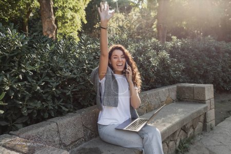 Fröhliche und lockige Frau winkt mit der Hand und spricht mit dem Smartphone, schaut weg und hält Laptop in der Hand, während sie tagsüber auf einer steinernen Bank im grünen Park in Barcelona sitzt 