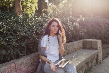 Junge, lockige Freiberuflerin in lässiger Kleidung, die auf einem Smartphone neben dem Laptop spricht, während sie auf einer steinernen Bank sitzt und Zeit in der Nähe grüner Pflanzen im Park in Barcelona, Spanien verbringt 