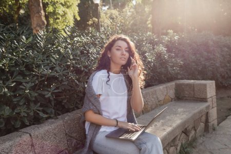 Junge und lockige Freiberuflerin in lässiger Kleidung, die mit dem Smartphone spricht und wegschaut, während sie Laptop benutzt und auf einer steinernen Bank im grünen Park in Barcelona sitzt, Spanien 