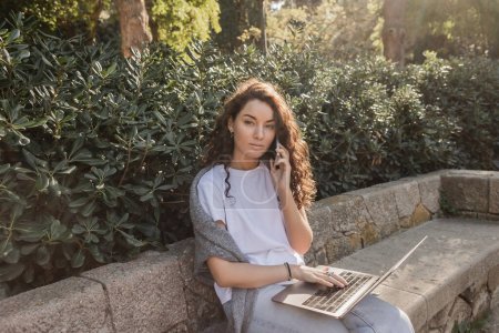 Young and curly freelancer in casual clothes talking on smartphone and using laptop while looking at camera on stone bench near green bushes in park in Barcelona, Spain 