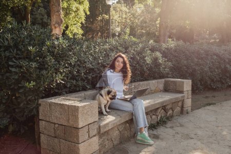 Young and curly freelancer looking at camera while using devices and petting pug dog and sitting on stone bench beside green plants in park in Barcelona, Spain 