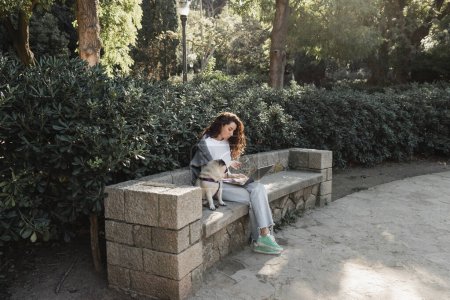 Lockige Freiberuflerin in lässiger Kleidung mit Gadgets, während sie tagsüber in Barcelona auf einer steinernen Bank im grünen Park sitzt und in der Nähe eines Mops arbeitet 