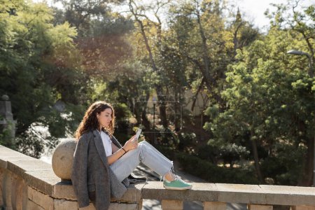 Junge und lockige Frau in warmer Jacke und lässiger Kleidung mit Smartphone und kabelgebundenen Kopfhörern, während sie am Zaun sitzt und Zeit im verschwommenen Park in Barcelona, Spanien verbringt 