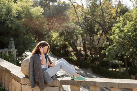 Young and curly freelancer in warm jacket using smartphone and earphones while holding laptop and sitting on stone fence in blurred summer park in Barcelona, Spain 