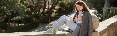 Joven freelancer rizado con chaqueta abrigada usando un smartphone con auriculares y sosteniendo un portátil mientras está sentado en un banco de piedra en un parque borroso en Barcelona, España, pancarta 