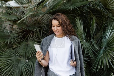 Fröhliche junge und lockige Freiberuflerin in warmer Jacke mit Kopfhörer und Smartphone, während sie mit Laptop neben grünen Palmen im Park in Barcelona steht, Spanien 