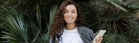 Positive and young curly freelancer in earphones and casual clothes holding gadgets and looking at camera while standing near palm trees in park at daytime in Barcelona, Spain, banner 