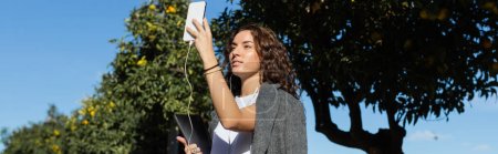 Young curly freelancer in warm jacket using smartphone and earphones while holding laptop and standing near green trees at background in park at daytime in Barcelona, Spain, banner 