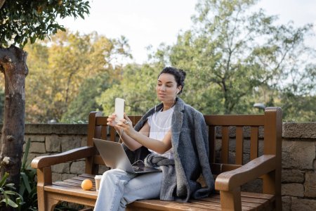 Joven freelancer despreocupado en auriculares usando teléfono celular y sosteniendo el portátil mientras está sentado cerca de naranja y café para ir a un banco de madera en el parque en Barcelona, España 